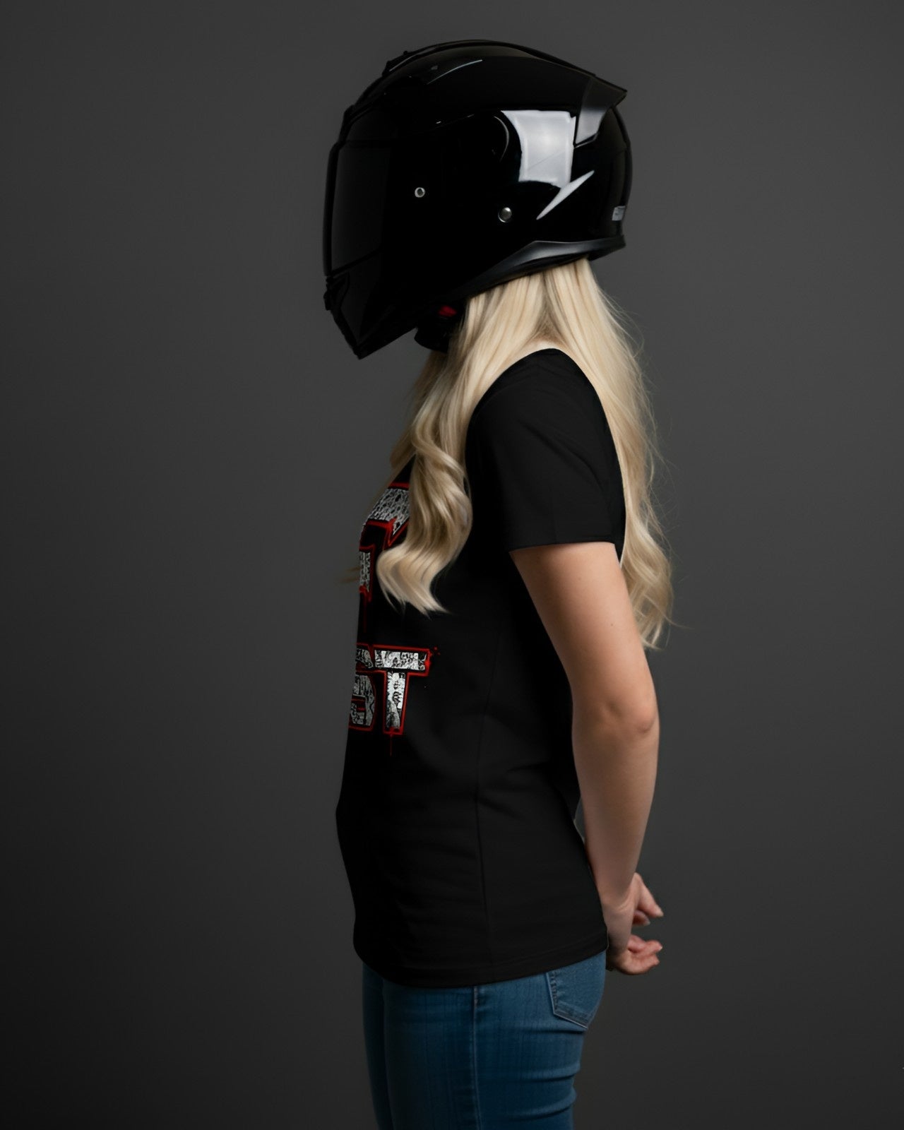 A woman with blonde hair, wearing a black superbike t-shirt and motorcycle enthusiast apparel, stands with a helmet on her head.