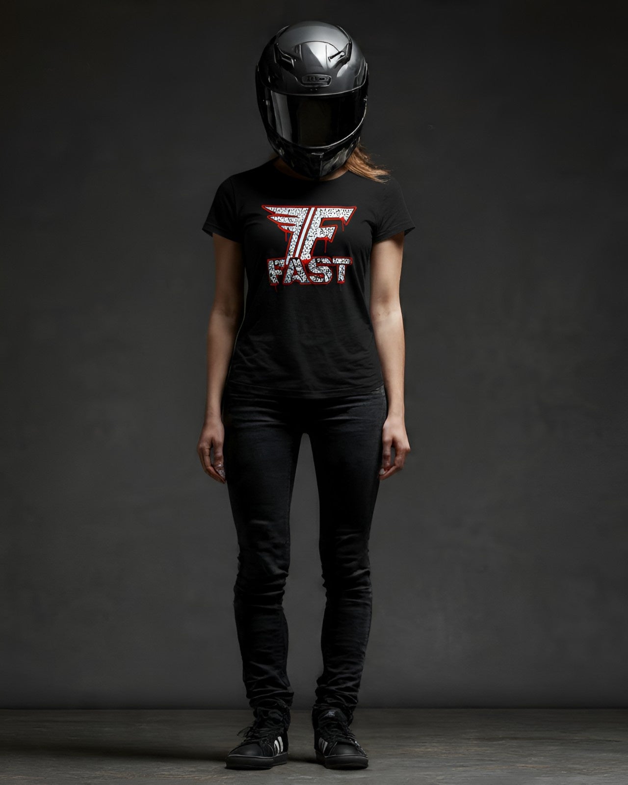 A full-body studio shot of a woman wearing a superbike t-shirt, motorcycle enthusiast apparel, and art shirt, black pants, and a black helmet.