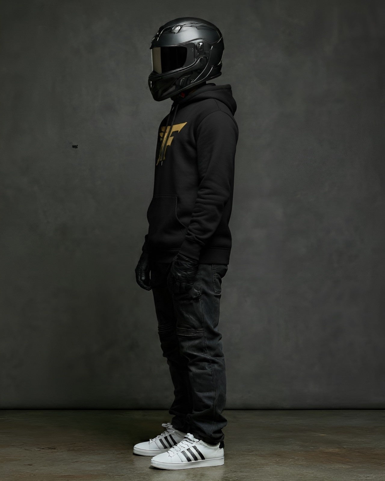 Person wearing a black helmet, black hoodie with a gold 'F' logo on the chest, black pants, and white sneakers against a dark background