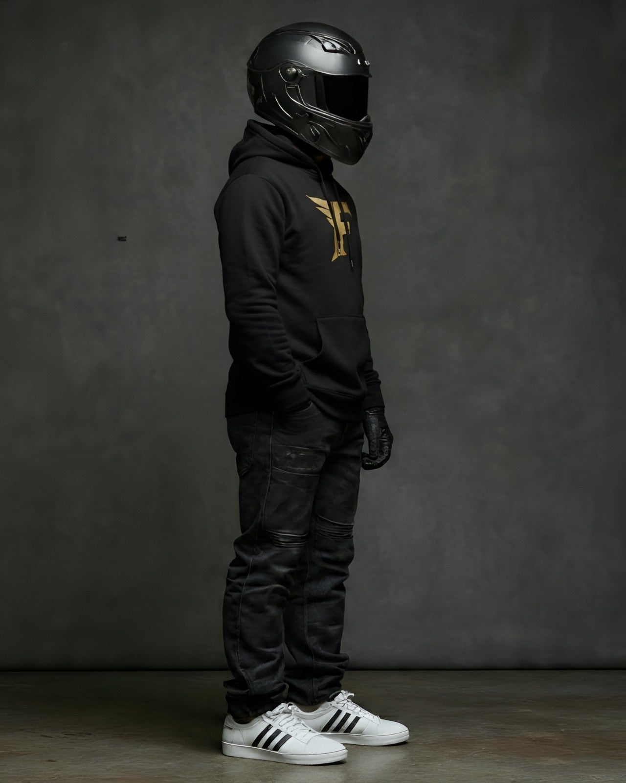 Person wearing a black helmet, black hoodie with a gold 'F' logo, black pants, and white sneakers on a gray background