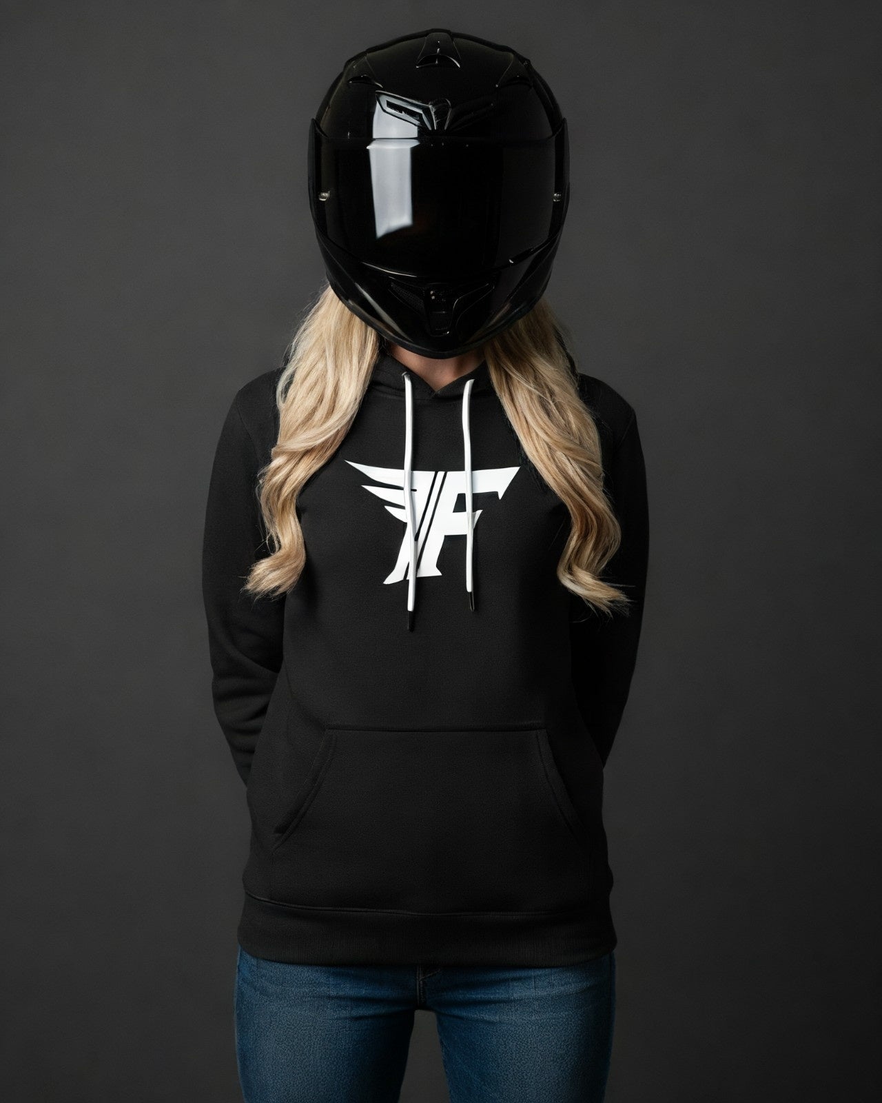 A blonde woman wearing a black motorcycle helmet and a black hoodie with a white "F" logo on the chest stands against a gray background.