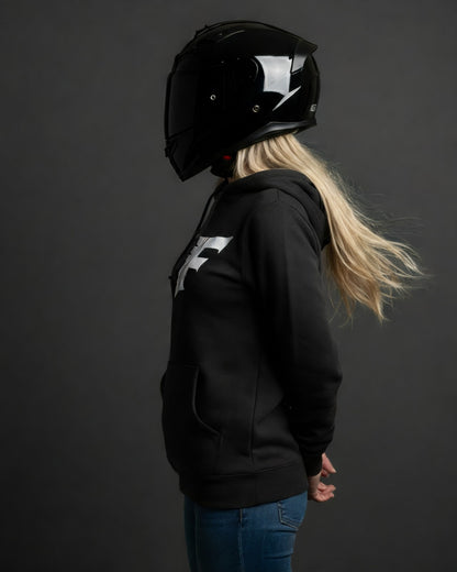 Blonde woman in a black, pullover hoodie with a white logo on the chest, wearing a glossy black motorcycle helmet, viewed from the left side against a grey background.