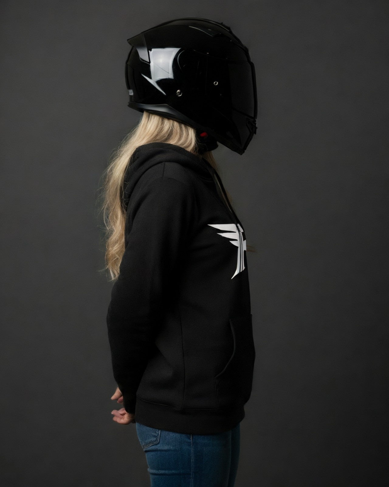 A blonde woman wears a black motorcycle helmet, a black hoodie with a white wing logo (the shirt), and blue jeans, standing in profile against a grey background.