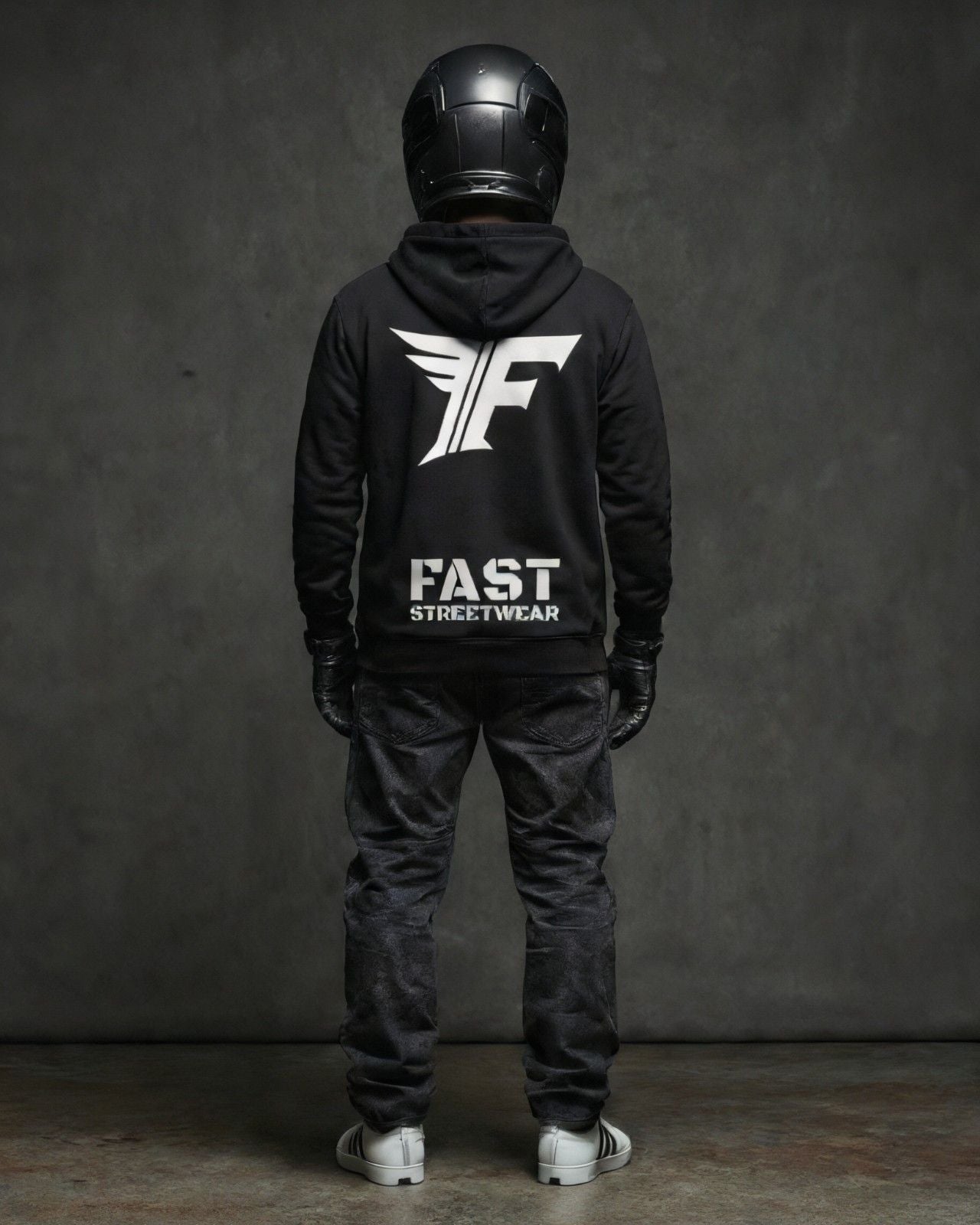 A person in a black hoodie, black jeans, and helmet stands facing away. The hoodie features a large white "F" logo with wings above the words "FAST STREETWEAR."