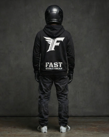 A person in a black hoodie, black jeans, and helmet stands facing away. The hoodie features a large white "F" logo with wings above the words "FAST STREETWEAR."