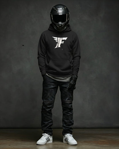 Man in a black pullover hoodie with a white winged 'F' logo, black textured jeans, motorcycle gloves, and white sneakers, standing against a dark grey background.