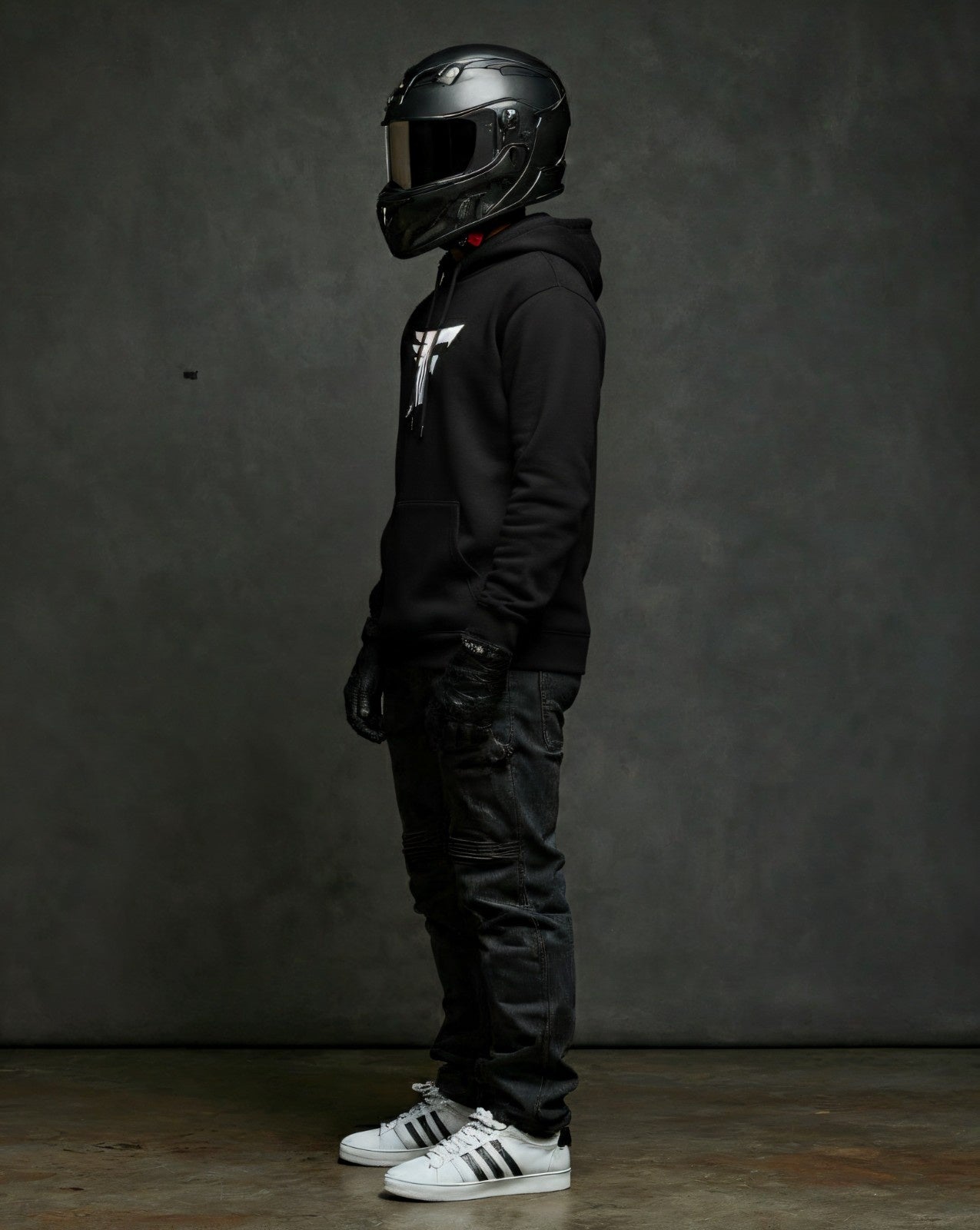 Studio photo of a person standing, wearing a black helmet, dark jeans, and black gloves. The focus is on their black hooded sweatshirt, which has a distinct white logo on the front.