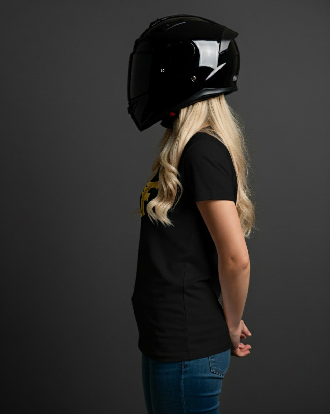 Black t-shirt on a blonde woman in a full-face motorcycle helmet, standing in profile against a dark background, hands claspedbehind her back.