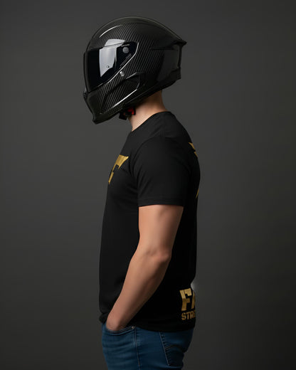 A person in profile wearing a carbon fiber motorcycle helmet and a black t-shirt with a gold graphic logo on the chest and lower torso.