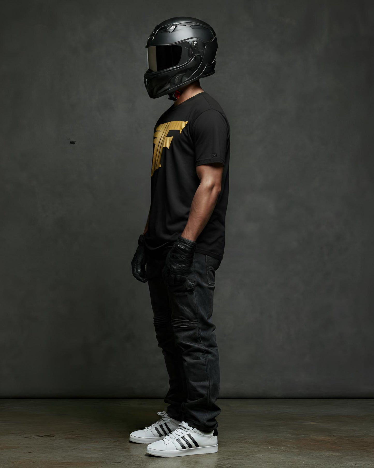A man, wearing a helmet, gloves, and black jeans, stands sideways, showcasing a black t-shirt with a bold gold graphic logo on the chest.