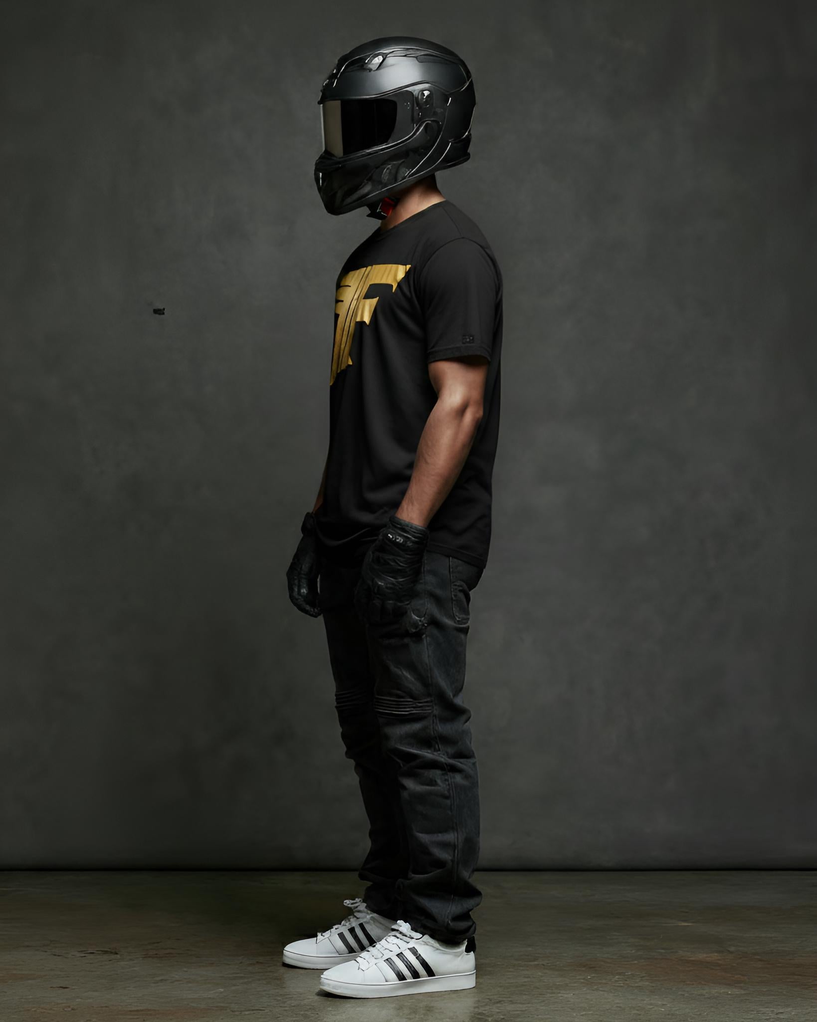 A man, wearing a helmet, gloves, and black jeans, stands sideways, showcasing a black t-shirt with a bold gold graphic logo on the chest.
