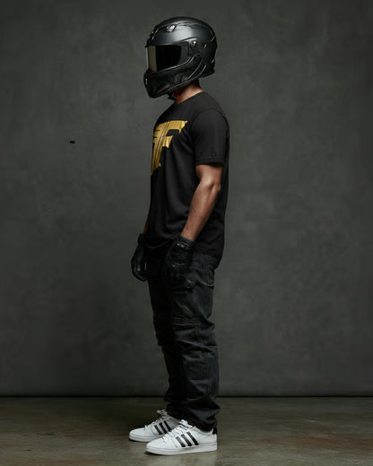 A man, wearing a helmet, gloves, and black jeans, stands sideways, showcasing a black t-shirt with a bold gold graphic logo on the chest.