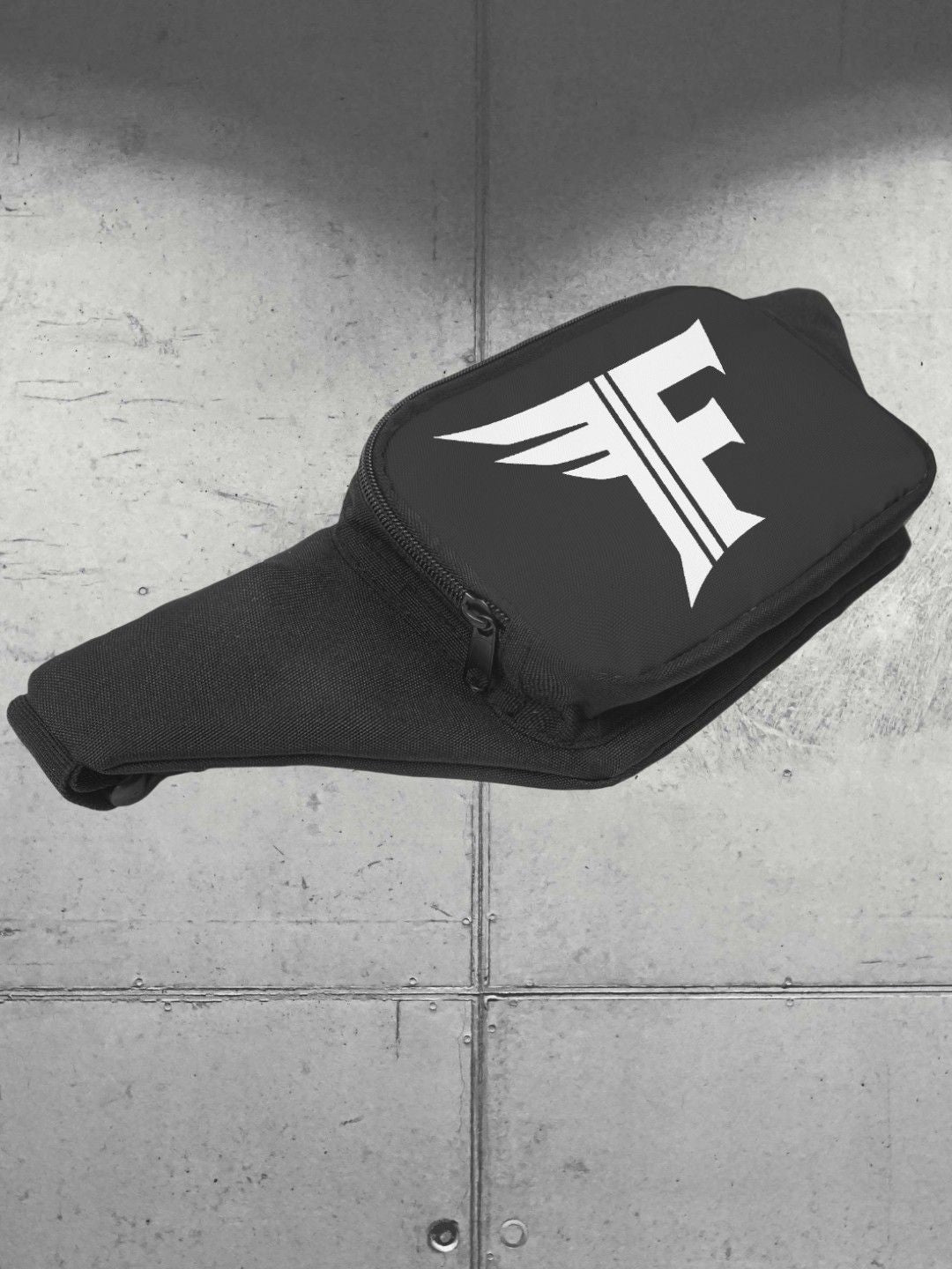 Black, durable fabric waist bag with zipper closure and a prominent white 'F' logo. Shot against a gray, industrial background.
