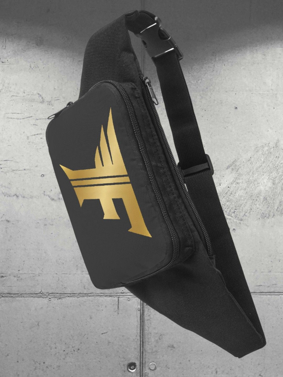 Close-up of a black sling/waist bag with a gold winged 'F' logo on the front pocket, hanging against a grey concrete wall.