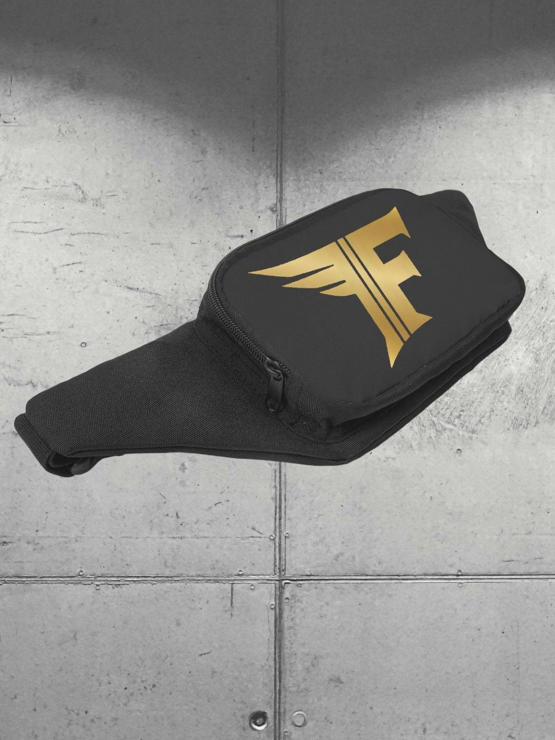 Angle view of the black waist bag (fanny pack) with a gold winged 'F' logo on the front pocket, set against a grey metal wall.