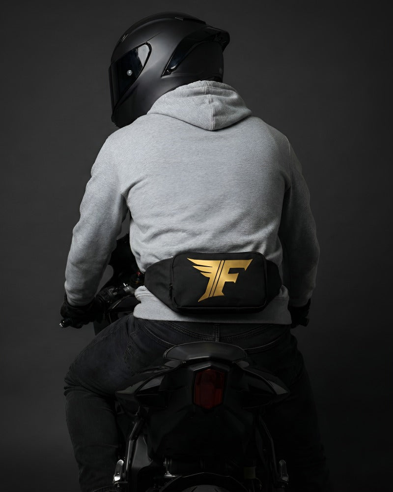 Black waist bag with gold winged 'F' logo worn on the back of a person in a grey hoodie and helmet riding a motorcycle.