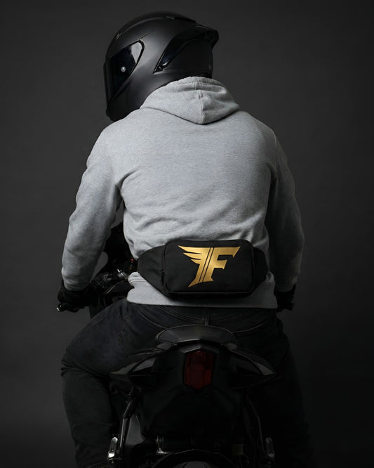 Black waist bag with gold winged 'F' logo worn on the back of a person in a grey hoodie and helmet riding a motorcycle.