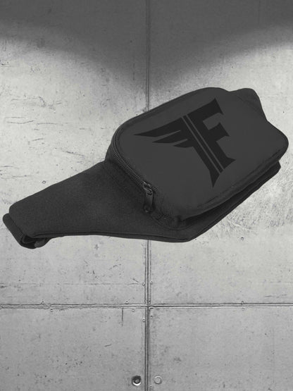 A close-up of a black fanny pack featuring a dark, stylized "F" logo on the main pouch, resting on a background of gray concrete panels.