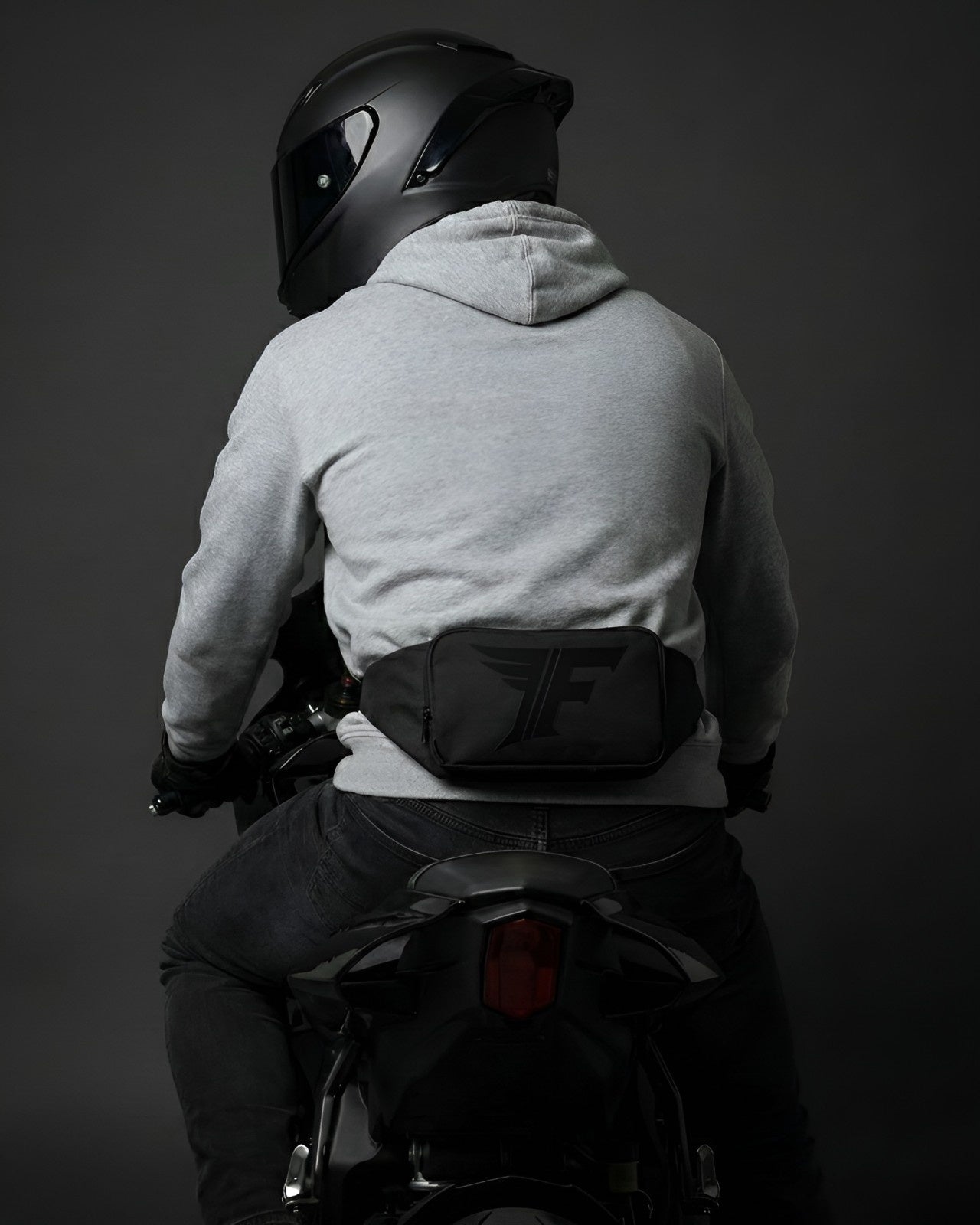 A person in a helmet and gray hoodie rides a motorcycle, wearing a black fanny pack with a logo prominently on their lower back.