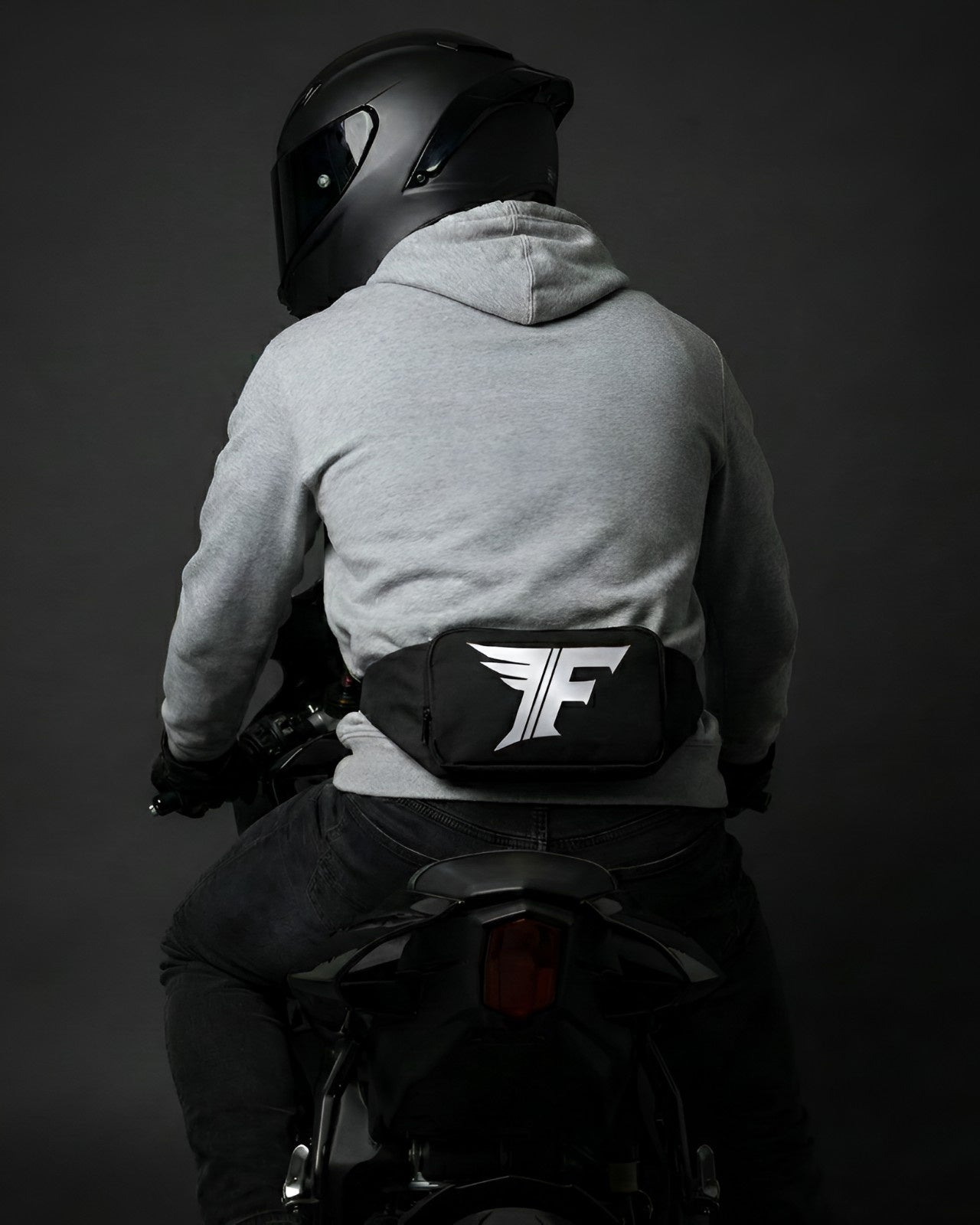 Close-up view of a black waist bag featuring a distinctive white winged 'F' logo, worn on the lower back of a helmeted motorcyclist.