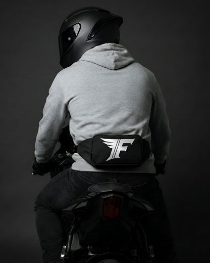 Close-up view of a black waist bag featuring a distinctive white winged 'F' logo, worn on the lower back of a helmeted motorcyclist.