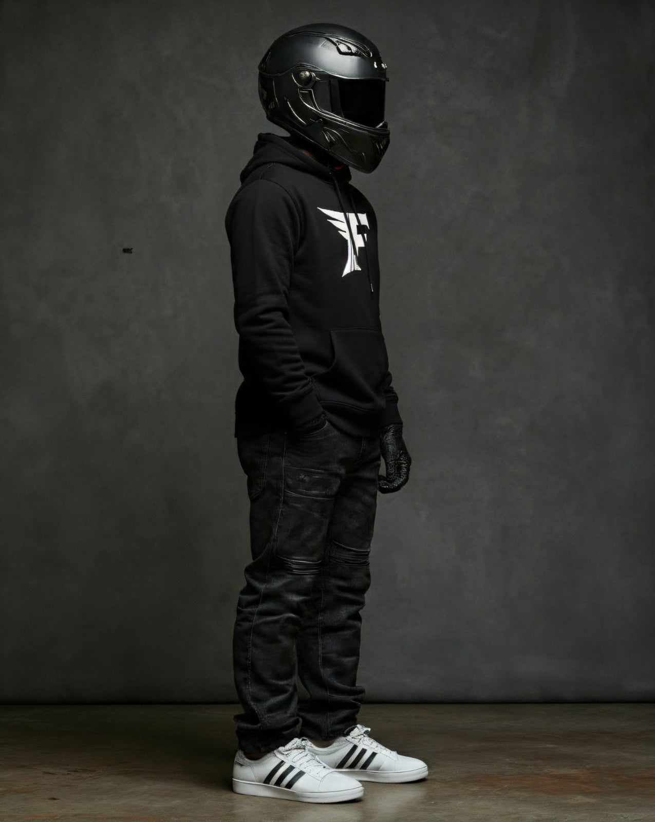 A person in a black motorcycle helmet, black hoodie with a white logo, dark jeans, and white Adidas sneakers stands in a full-right profile.