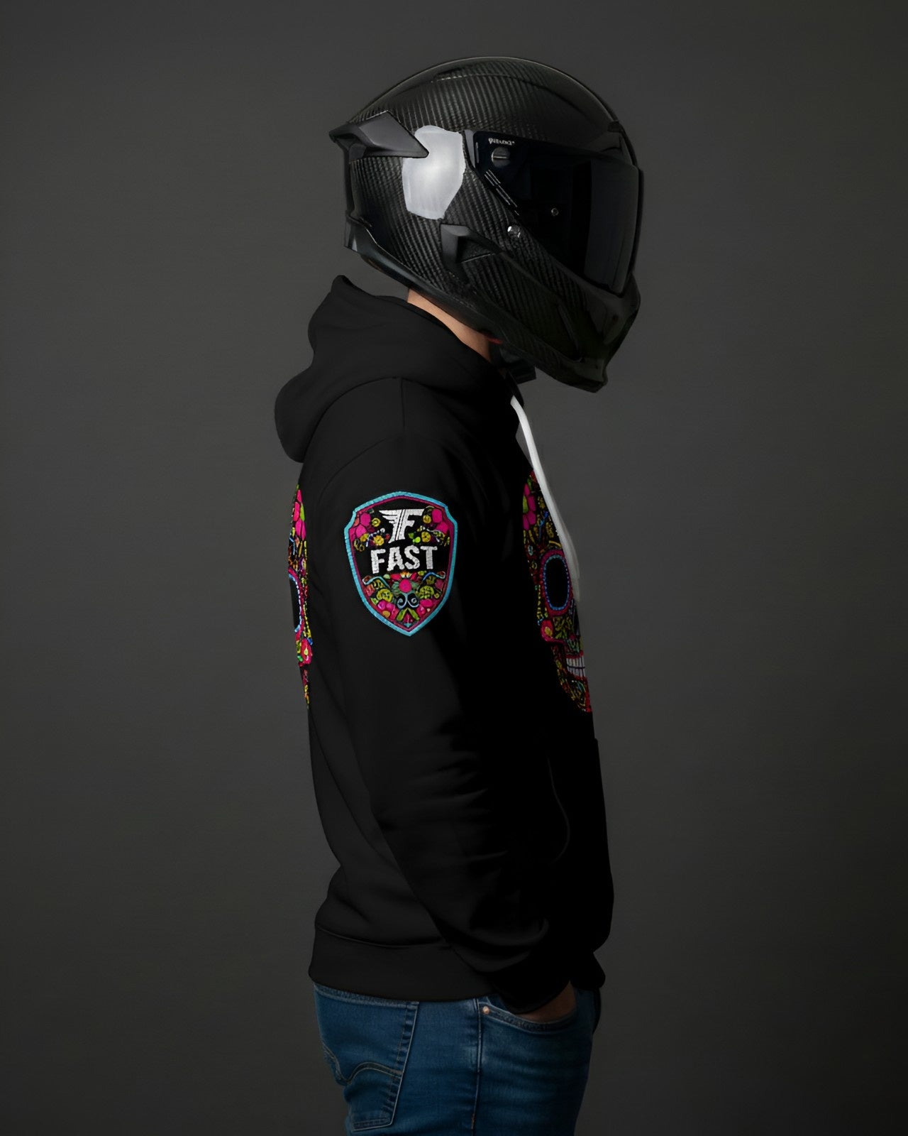 Right side view of black hoodie showing the colorful shield "FAST" logo patch on the right shoulder. Model wears a black motorcycle helmet.