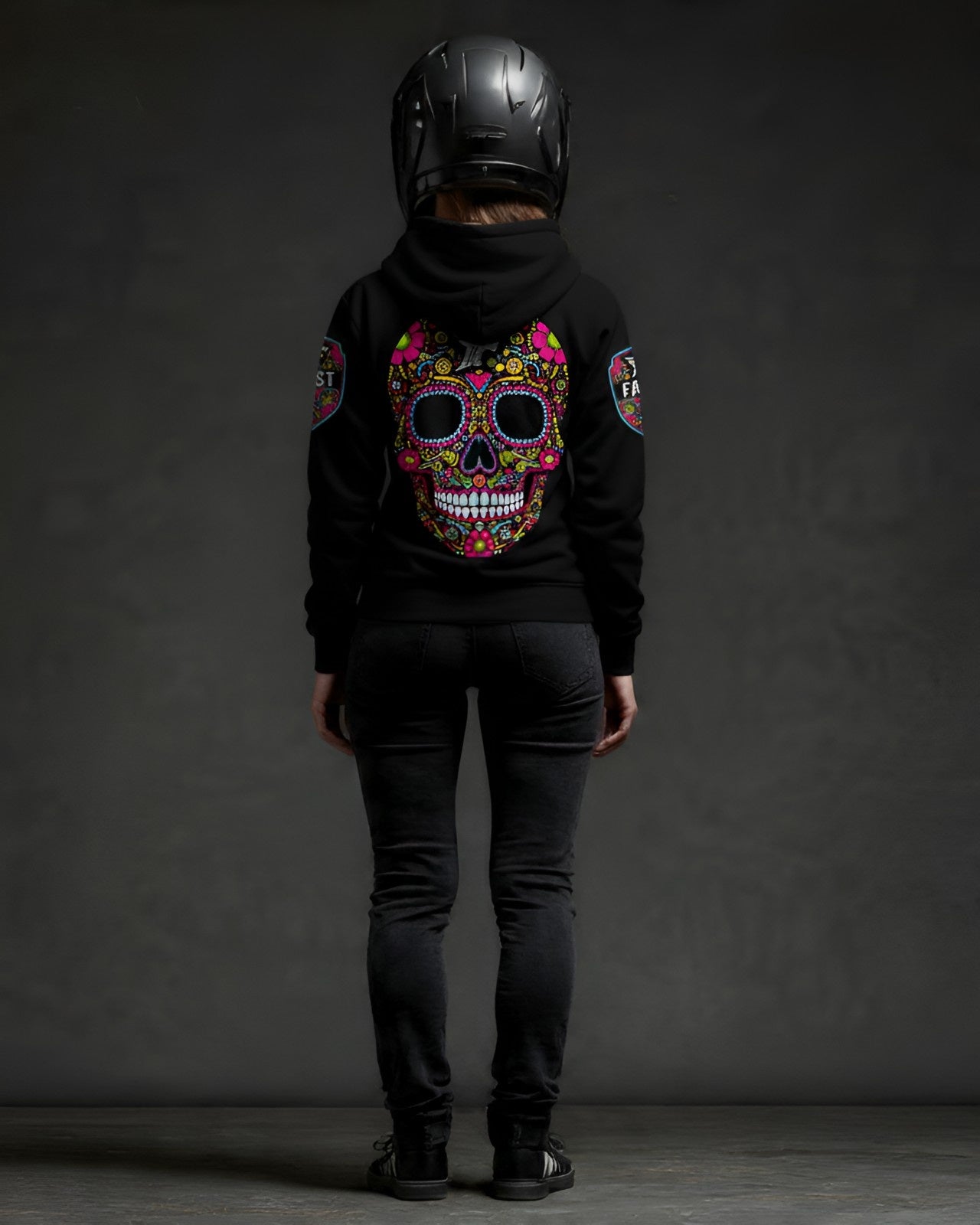 Back view of a black hoodie with large, colorful sugar skull design, worn by a figure in a motorcycle helmet. Shows sleeve patches and dark jeans.