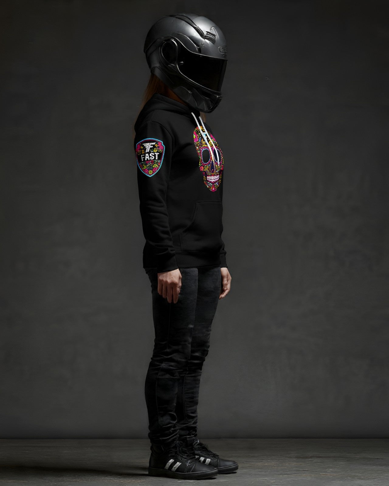 Right side profile of a black hoodie with a sugar skull design and a prominent sleeve patch, worn by a figure in a full-face motorcycle helmet.