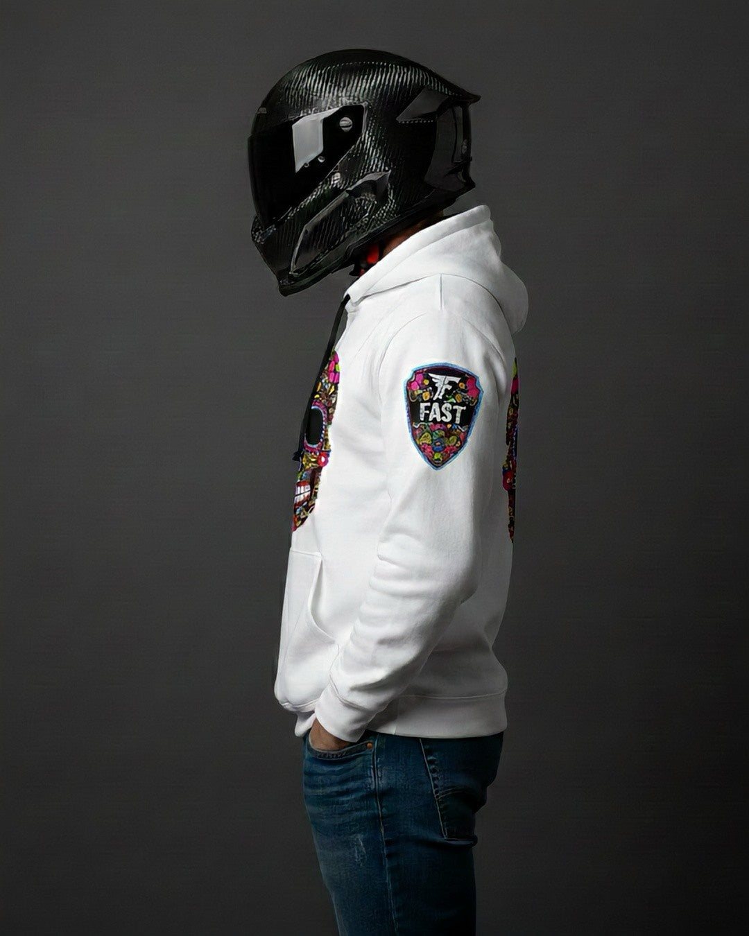 Left profile of the white sugar skull hoodie, showcasing the shield "FAST" logo patch on the left sleeve. Worn by a person in a motorcycle helmet.