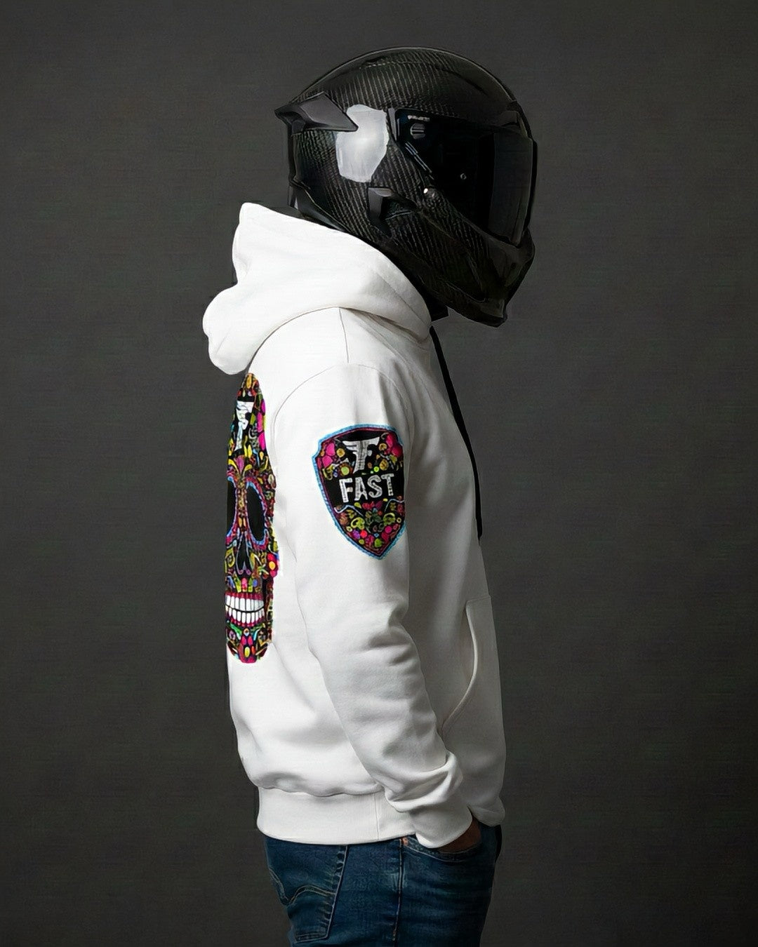 Right profile of the white sugar skull hoodie, showing the shield "FAST" logo patch on the right sleeve. Worn by a person in a motorcycle helmet.