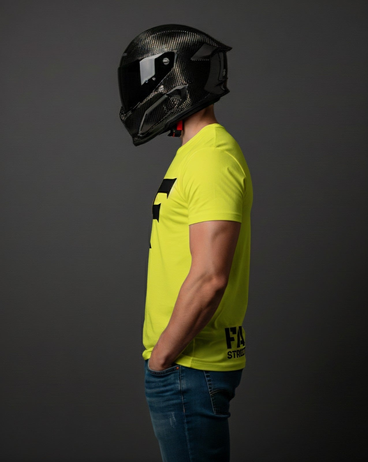 Profile of a person in a bright yellow t-shirt and ripped blue jeans, wearing a dark carbon fiber motorcycle helmet, hands in pockets.