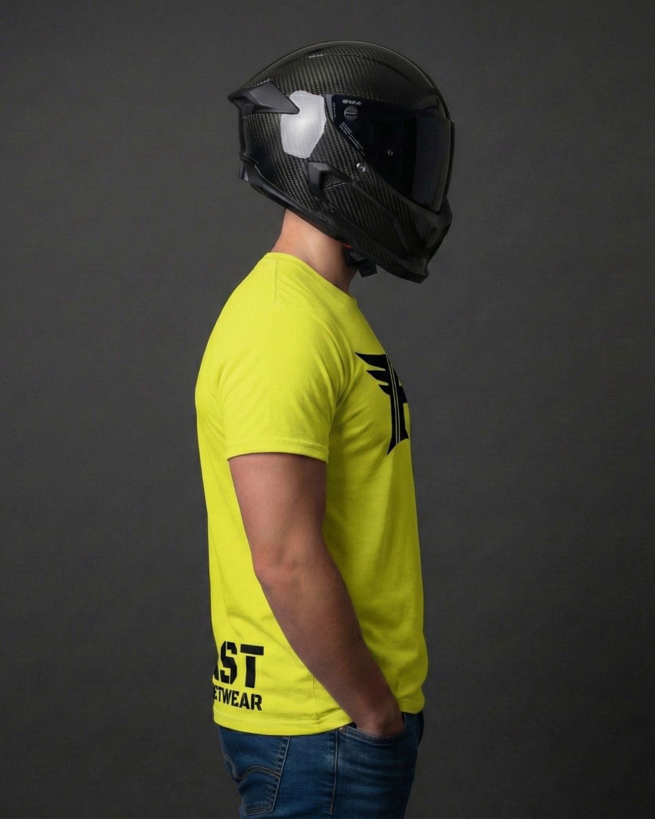 Man in a bright yellow t-shirt with a black graphic and text, hands in blue jeans pockets, wearing a black carbon fiber full-face motorcycle helmet.