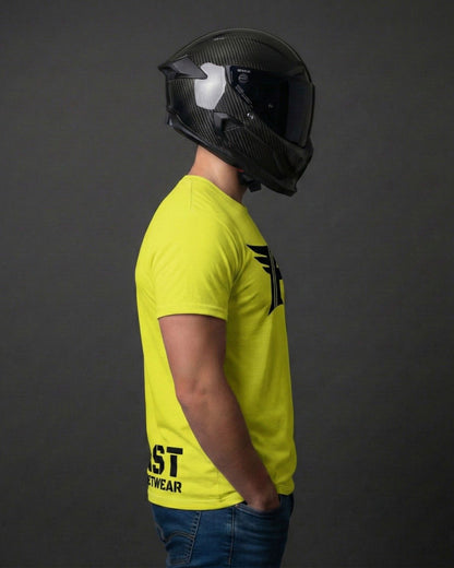 Man in a bright yellow t-shirt with a black graphic and text, hands in blue jeans pockets, wearing a black carbon fiber full-face motorcycle helmet.